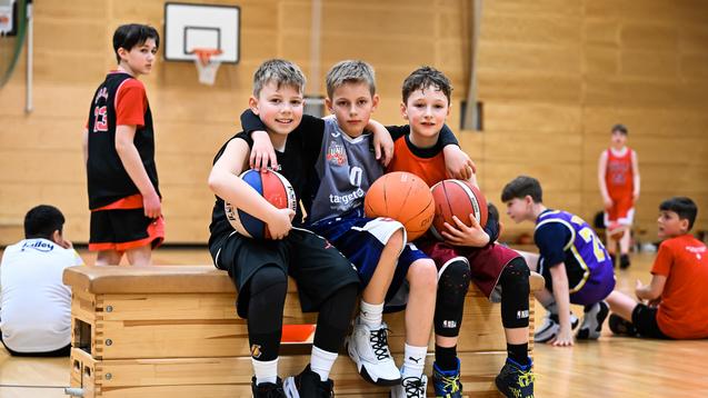 Drei Kinder posieren auf einer Bank in einer Sporthalle, jeder hält einen Basketball. Im Hintergrund weitere Spieler.