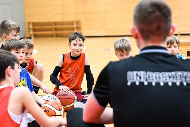 Gruppe von Jungen beim Basketballtraining, sitzend auf dem Boden mit Bällen, Trainer spricht zu ihnen.