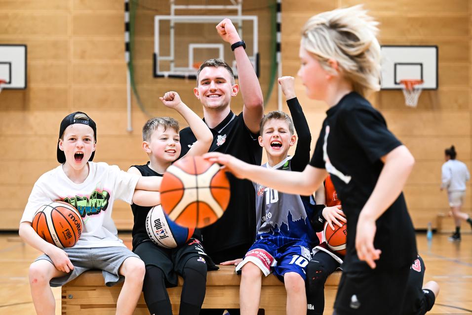 Gruppe fröhlicher Kinder und Trainer in Basketballkleidung posieren auf einer Bank in einer Sporthalle, mit Basketballs in den Händen.