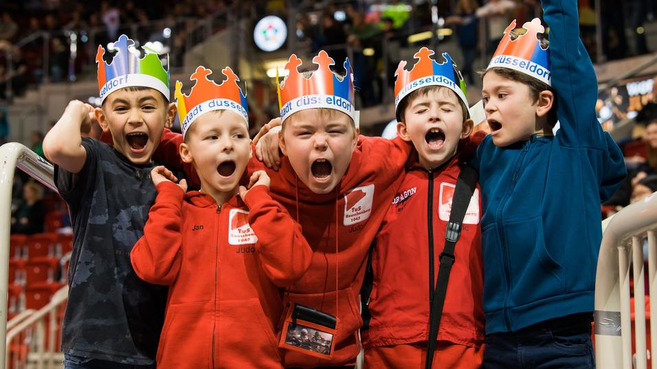 Gruppe von fünf jubelnden Kindern mit farbigen Kronen und roten Sweatshirts in einer Sportarena.