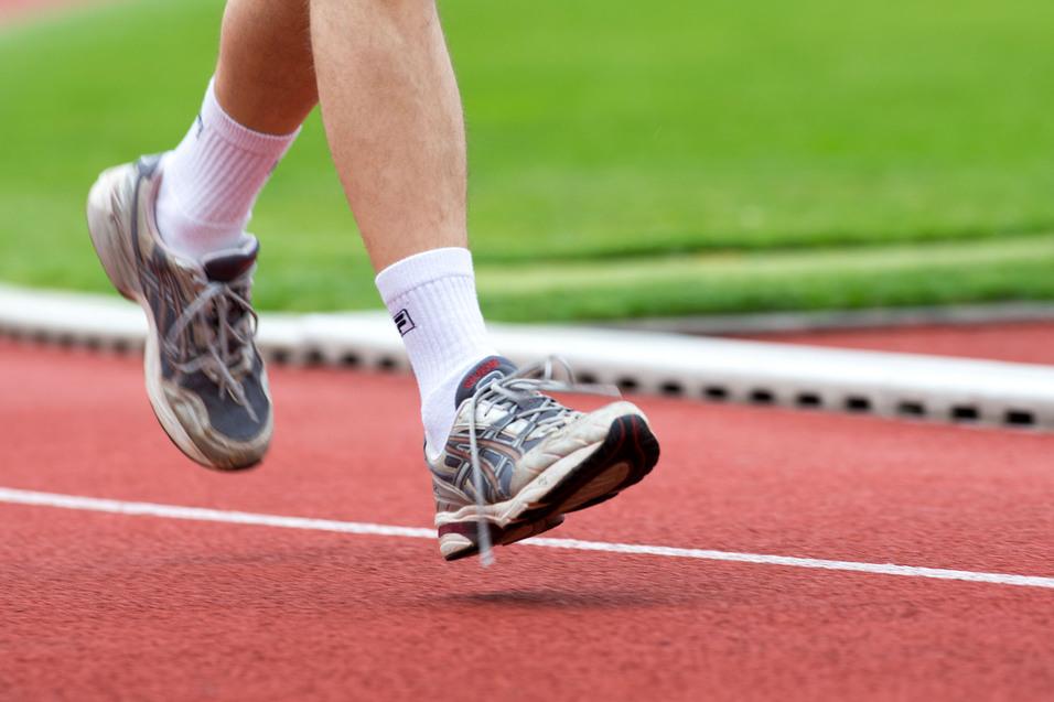 Läufer in grauen Sportschuhen hebt den Fuß auf einer roten Laufbahn über grünem Rasen.