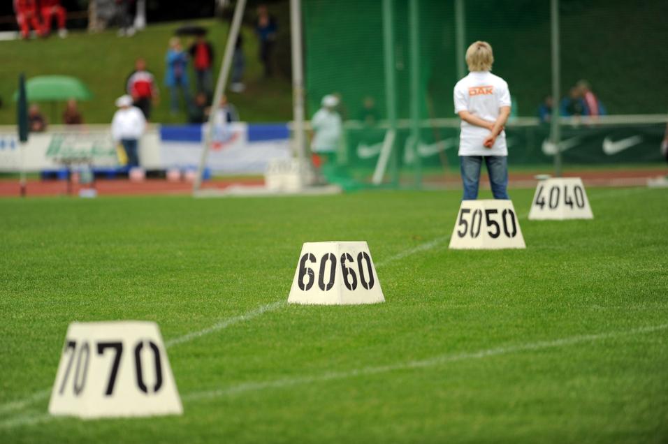 Person mit kurzen Haaren steht auf einem Sportplatz vor nummerierten Markierungen (40, 50, 60, 70) im Gras.