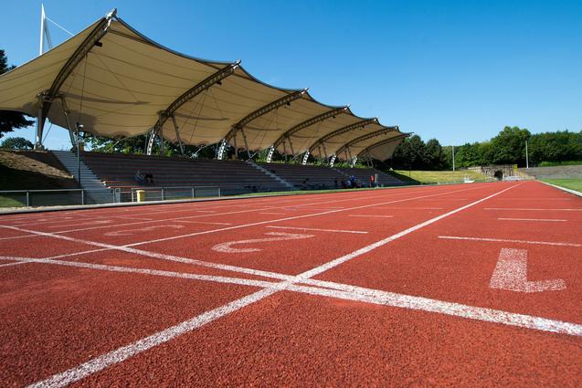 Nahaufnahme einer roten Laufbahn mit Startlinien und einem überdachten Zuschauerbereich im Hintergrund.