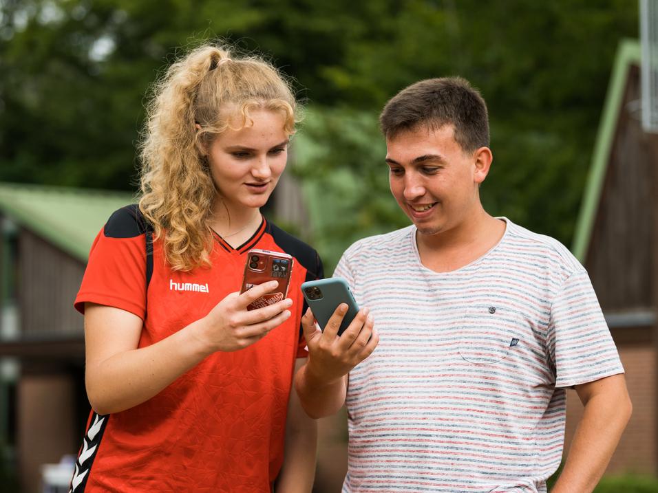 Zwei junge Leute schauen auf ihre Smartphones, stehen lächelnd im Freien vor einem Gebäude.