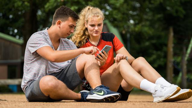 Zwei Jugendliche sitzen auf dem Boden und betrachten gemeinsam ein Smartphone. Ein Mädchen trägt ein rotes sportliches Oberteil.