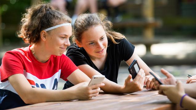Zwei Mädchen sitzen an einem Tisch, schauen auf ihre Smartphones und lächeln dabei. Im Hintergrund sind weitere Personen sichtbar.