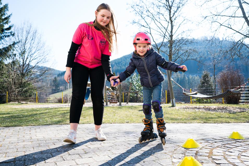 Ein Mädchen übt mit einer Trainerin das Inline-Skaten auf einem gepflasterten Platz, umgeben von Bäumen und Hügeln.