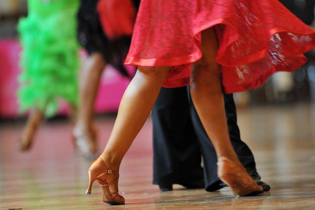 Tanzende Beine in eleganten Schuhen, während sich ein rotes Kleid dreht, vor unscharfem Hintergrund.