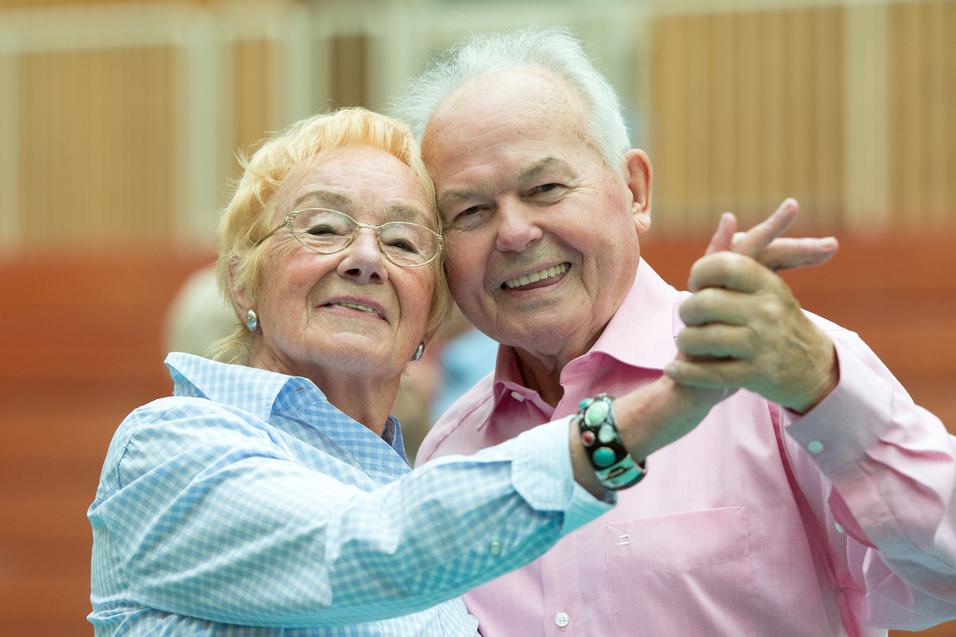 Ein älteres Paar umarmt sich lächelnd und tanzt in einer Sporthalle. Ihre Kleidung ist hell und farbenfroh.