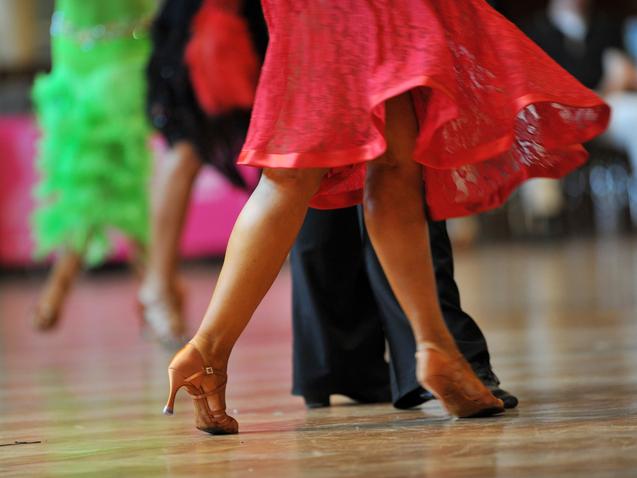 Tanzende Beine in eleganten Schuhen, während sich ein rotes Kleid dreht, vor unscharfem Hintergrund.