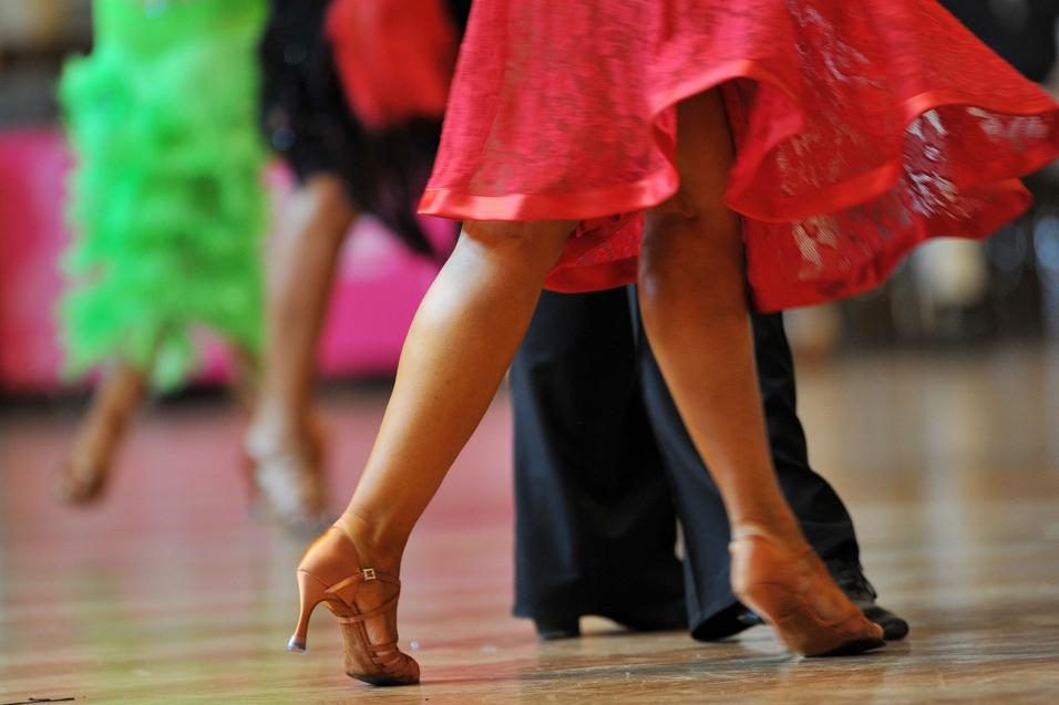 Tanzende Beine in eleganten Schuhen, während sich ein rotes Kleid dreht, vor unscharfem Hintergrund.