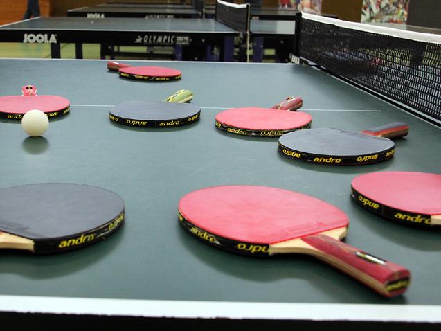 Mehrere Tischtennisschläger in verschiedenen Farben und ein Tischtennisball liegen auf einer grünen Tischtennisplatte.
