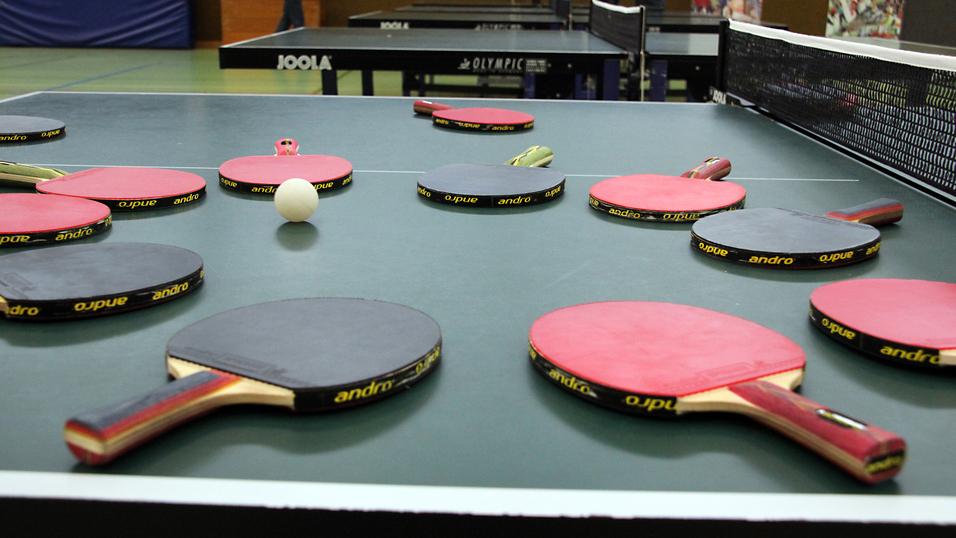 Mehrere Tischtennisschläger in verschiedenen Farben und ein Tischtennisball liegen auf einer grünen Tischtennisplatte.