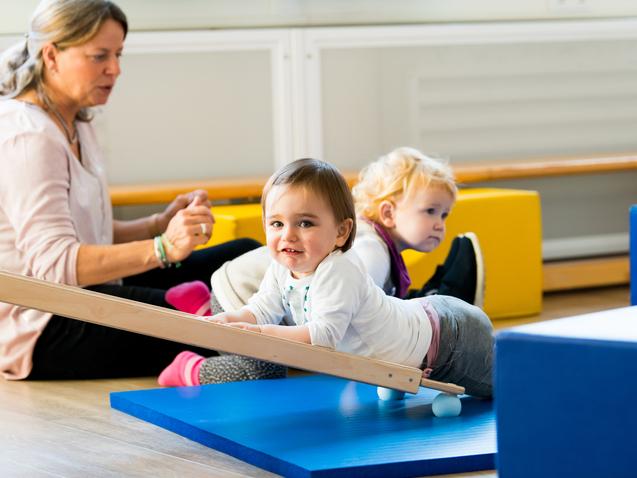 Zwei Kleinkinder spielen auf einer blauen Matte, während eine betreuende Person im Hintergrund sitzt.